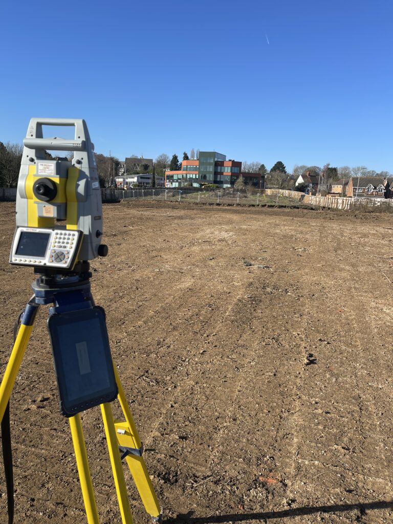 Image of a total station surveying instrument overlooking a muddy construction site with a large office building in the background, blue sky and sunny day.